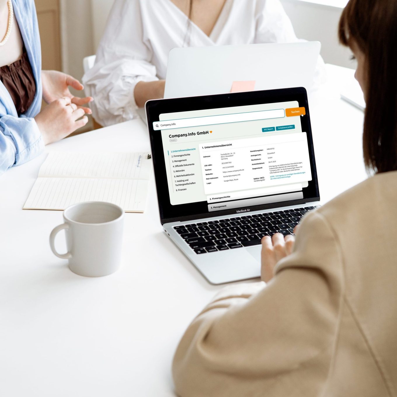 Three professionals in a bright meeting room reviewing the company.info company profile portal on a MacBook Air, showing the detailed company overview for "Company.Info GmbH" with sidebar navigation, KYC Report and Compliance check buttons visible on screen.