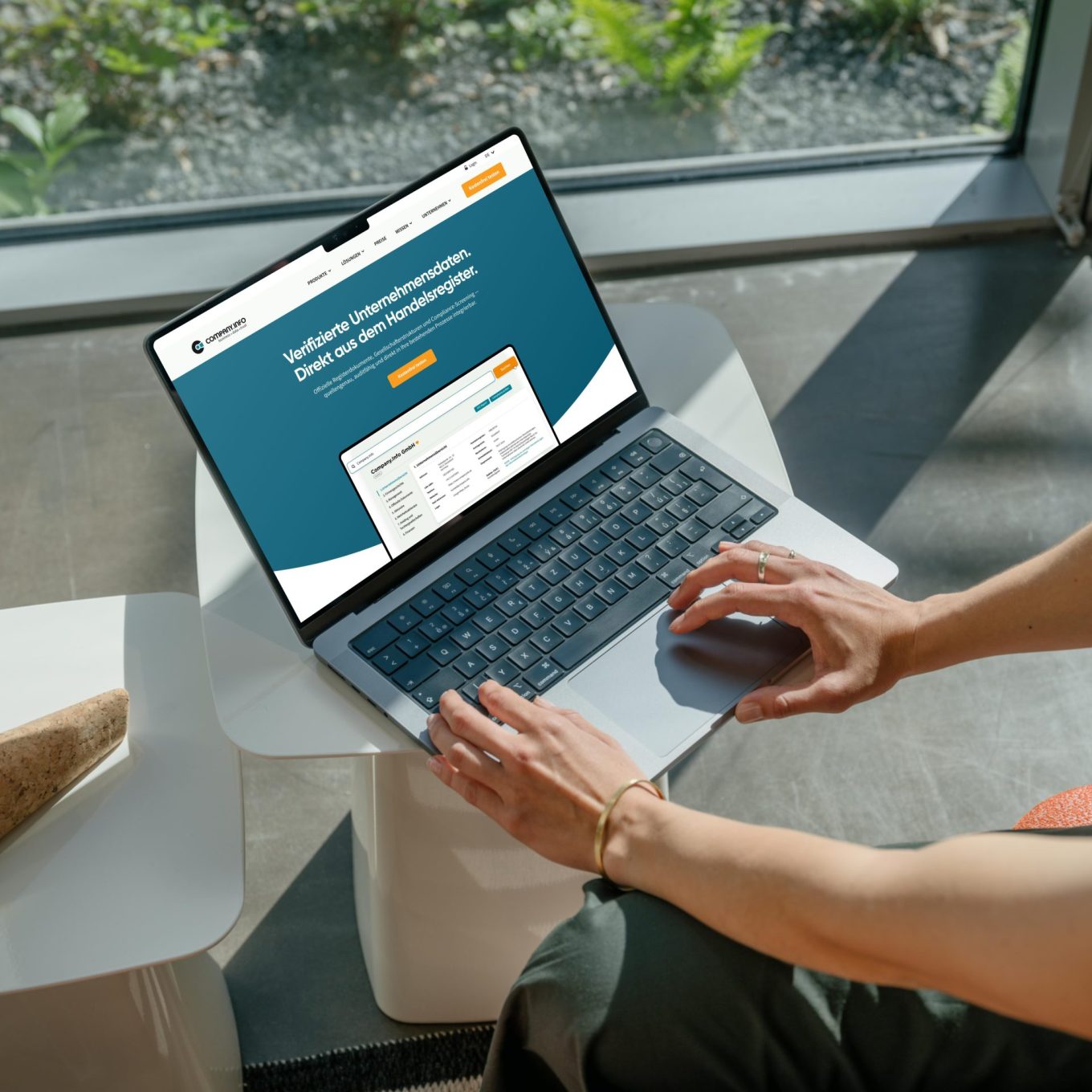 Woman working on a MacBook Pro in a bright modern office space, screen showing the company.info portal homepage with the headline "Verifizierte Unternehmensdaten. Direkt aus dem Handelsregister."