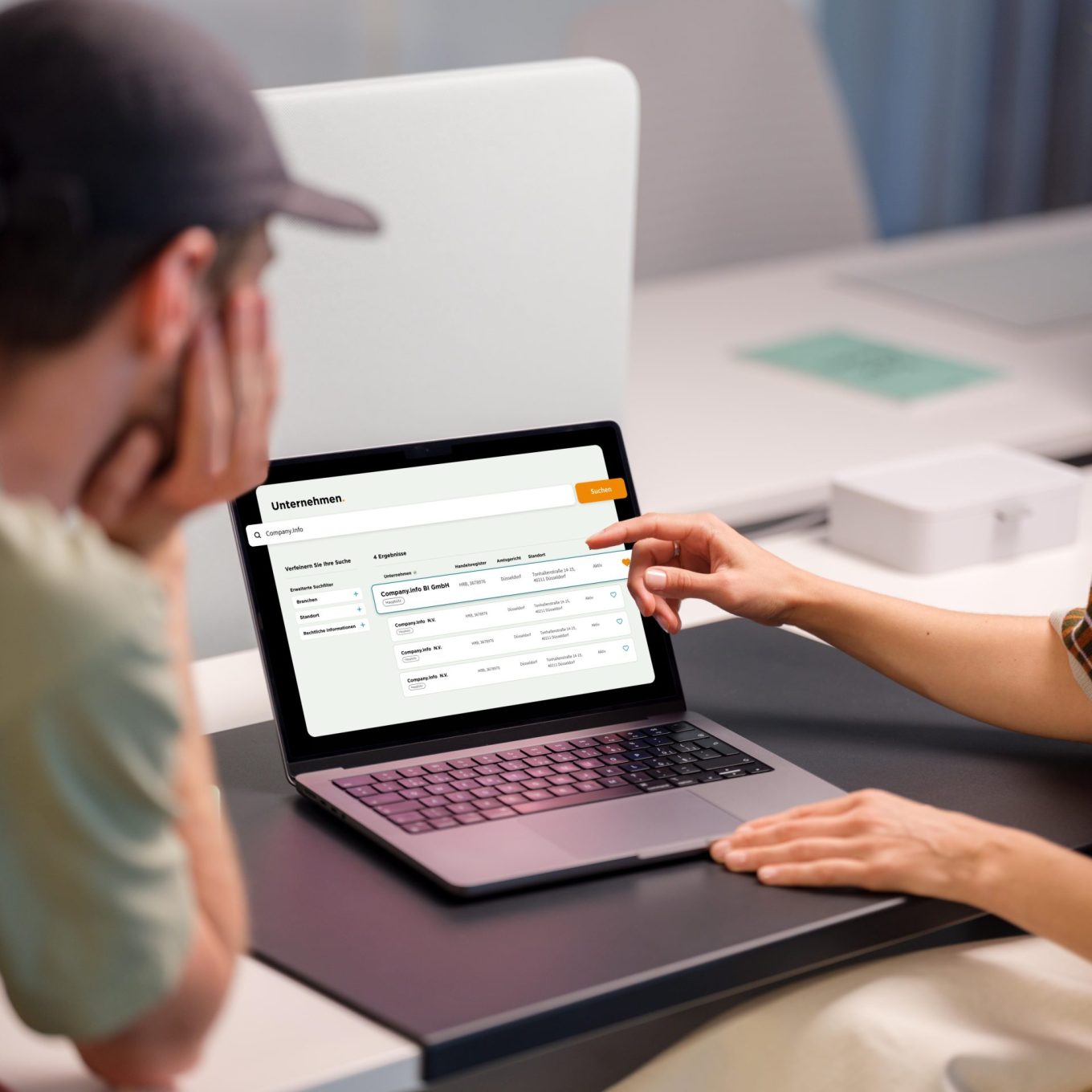 Two colleagues at a desk reviewing company search results on the company.info portal, with one person pointing at the screen showing filtered results including company name, commercial register number, court, location and status.